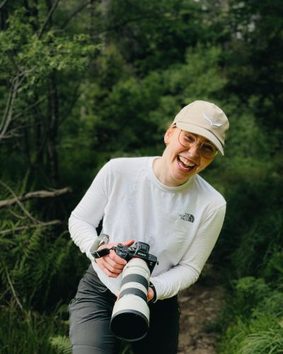 Eine Frau steht im Wald mit einer Kamera in der Hand und lacht.